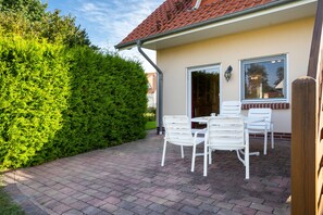 A patio with white chairs and a table, a neatly trimmed hedge, and a building with a red-tiled roof. - Hus Utblick in Fuhlendorf (Fuhlendorf)