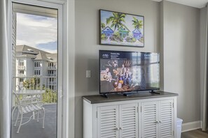 A living room with a flat-screen TV on a cabinet, a balcony with a table and chairs, and a painting of a beach scene. - Luxury Baytowne Wharf Condo with Beach and Pool (Miramar Beach)