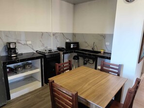A compact kitchen with a stove, coffee maker, and a small sink. There is a wooden table with chairs and a shelf with pots. - Cozy 2-Bedroom Apartment with AC - San Cristóbal Galápagos (Galápagos Province)