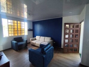 A room with a blue wall, a beige sofa, a blue armchair, and a wooden door with glass panels. - Cozy 2-Bedroom Apartment with AC - San Cristóbal Galápagos (Galápagos Province)