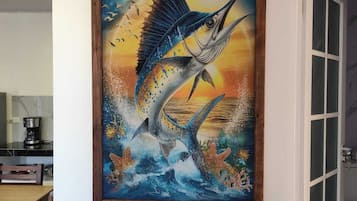 Una obra de arte enmarcada muestra un pez saltando sobre el agua, con un atardecer de fondo, montada en una pared blanca en una habitación con una puerta y una mesa con una cafetera.