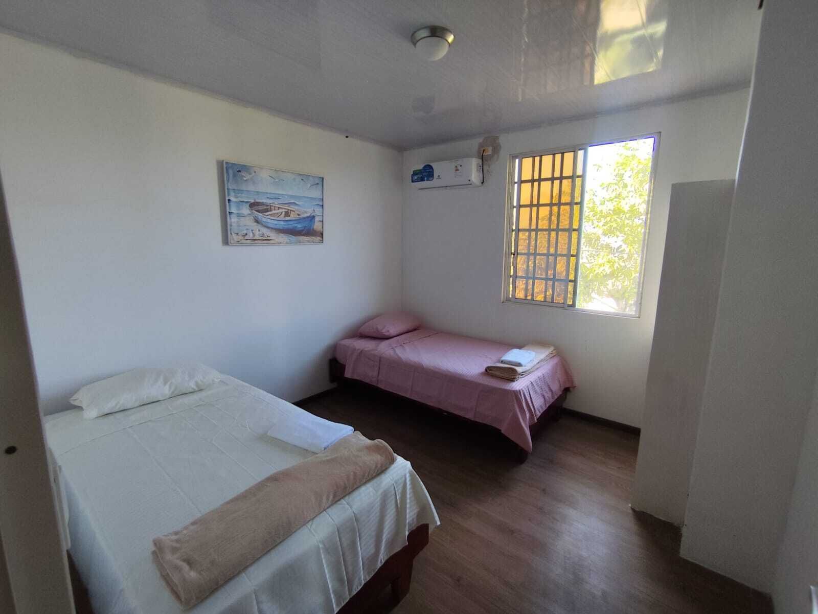 Una habitación con dos camas, una con cubrecolcha blanco y la otra con cubrecolcha rosa. Hay un cuadro en la pared y una ventana con barrotes.
