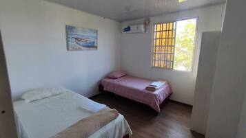 Una habitación con dos camas, una con cubrecolcha blanco y la otra con cubrecolcha rosa. Hay un cuadro en la pared y una ventana con barrotes.