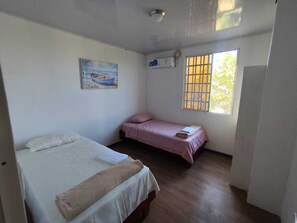 A room with two beds, one with a white bedspread and the other with a pink bedspread. There is a painting on the wall and a window with bars. - Cozy 2-Bedroom Apartment with AC - San Cristóbal Galápagos (Galápagos Province)