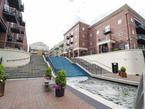Ein mehrstöckiges Gebäude mit Wasserspiel im Vordergrund, einer Treppe und einer Bank mit Blumentöpfen.