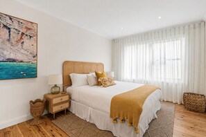 A bedroom with a bed, bedside table, lamp, and a painting on the wall. - Golden Sands The Best of Bondi on Your Doorstep (Bondi Beach)