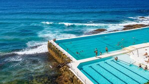 A large outdoor swimming pool with clear blue water, surrounded by rocky cliffs and the ocean. - Golden Sands The Best of Bondi on Your Doorstep (Bondi Beach)