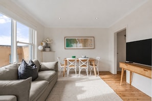 A modern living room with a sofa, dining table, chairs, a flat-screen TV, and a painting on the wall. - Golden Sands The Best of Bondi on Your Doorstep (Bondi Beach)