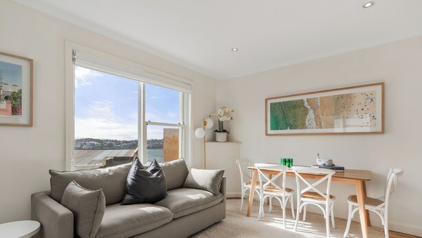 A modern living room with a sofa, a dining table with chairs, and a large window offering a city view. - Golden Sands The Best of Bondi on Your Doorstep (Bondi Beach)