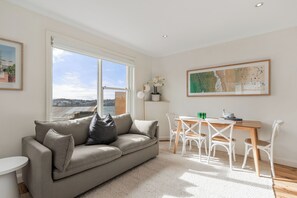 A modern living room with a sofa, a dining table with chairs, and a large window offering a city view. - Golden Sands The Best of Bondi on Your Doorstep (Bondi Beach)