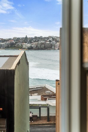 A coastal cityscape with buildings, a view of the sea, and a person standing on a sidewalk. - Golden Sands The Best of Bondi on Your Doorstep (Bondi Beach)