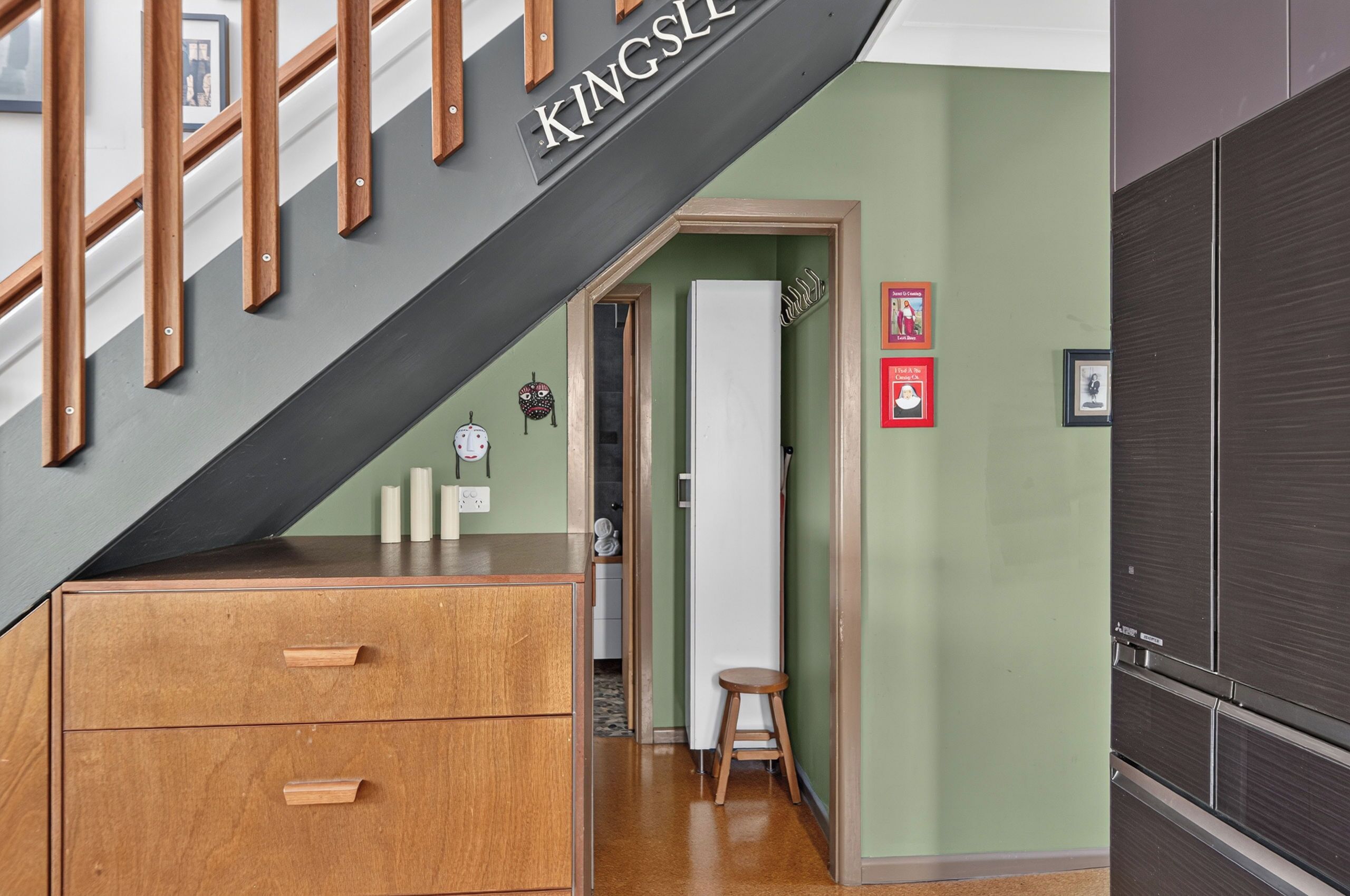 A staircase with a sign reading 'KINGSLEE' leading to a room with a wooden cabinet, a stool, and a wall-mounted shelf with framed pictures.
