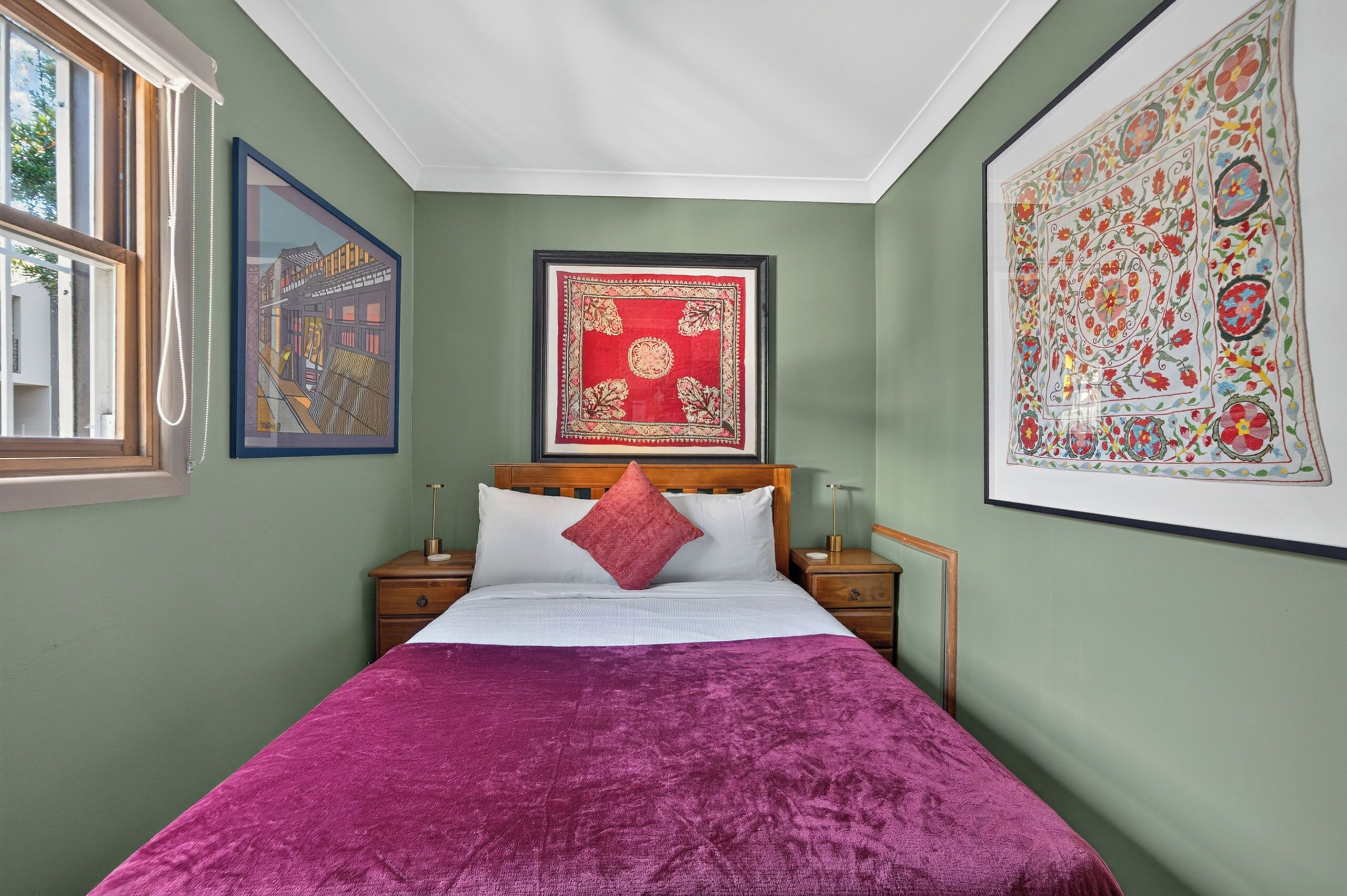 A room with a bed, two bedside tables, a window, and two framed artworks on the walls.