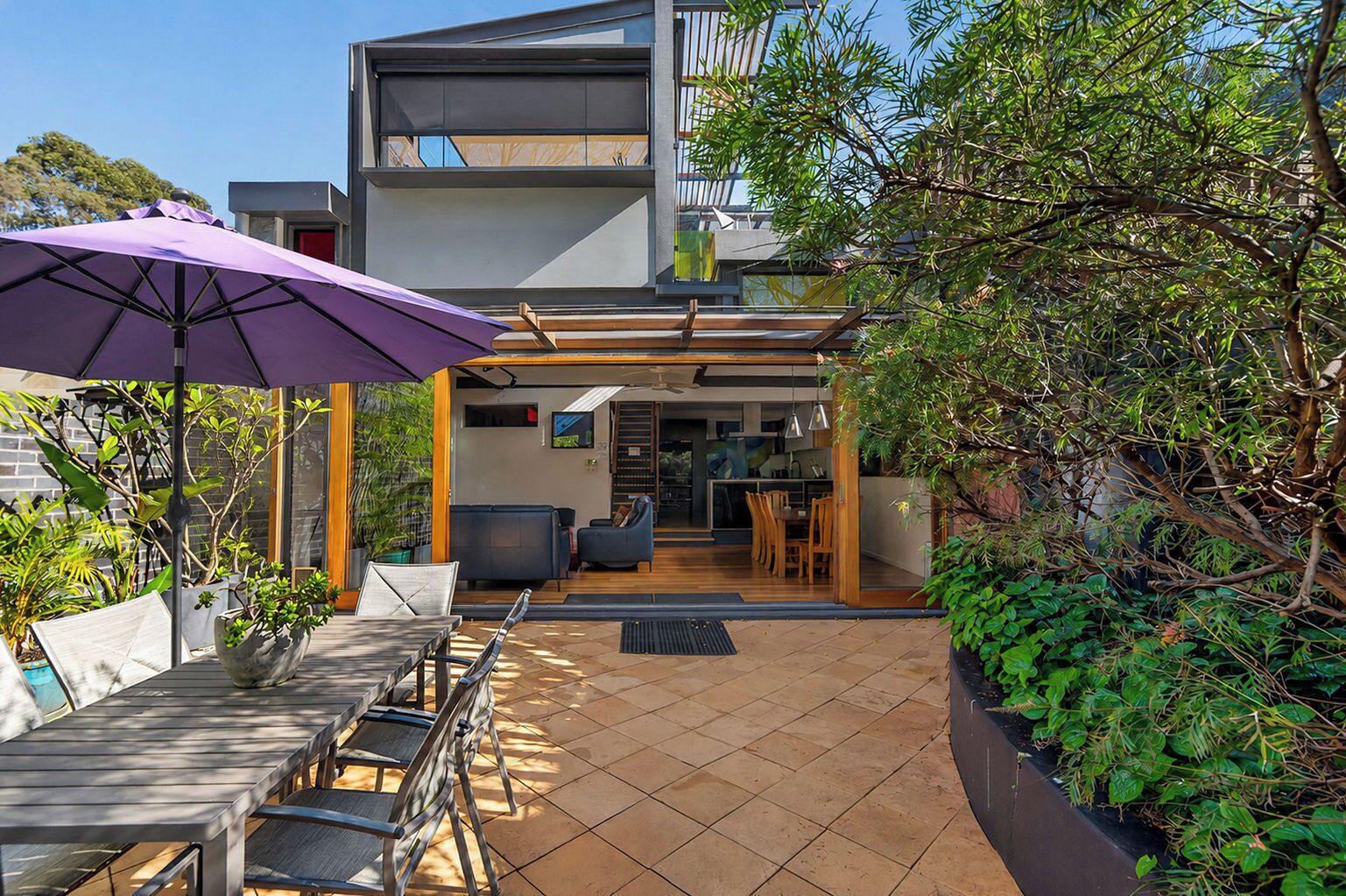 Une terrasse extérieure moderne, dotée d’un espace repas, d’un parasol violet et offrant une vue sur l’intérieur de la maison.