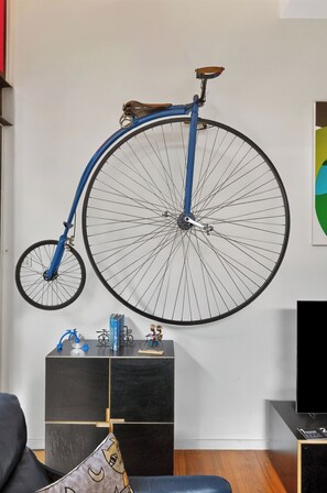 Ein Zimmer mit einem blauen Fahrrad an der Wand, einem schwarzen Sofa, einem kleinen Tisch mit Büchern und einem Fahrradmodell sowie einem Fernseher auf einem Standfuß.