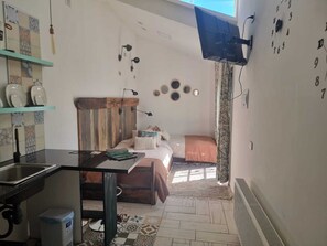 Ein kleines Zimmer mit einem Bett, einem Schreibtisch, einem kleinen Fenster und einer Wanduhr.