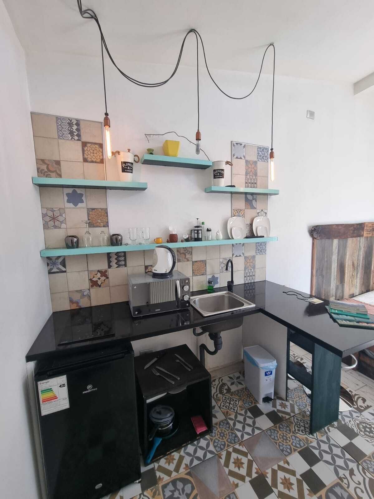 Een compacte keuken met een zwart aanrecht, een inbouwgootsteen en een kleine koelkast. Twee lampen hangen aan het plafond en de spatwand is voorzien van diverse tegels met verschillende patronen.
