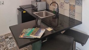 A compact kitchen with a black countertop, a sink, a toaster, and a small refrigerator. There are shelves above the countertop holding dishes and a yellow bowl.