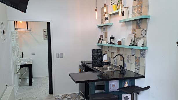 A compact kitchen with a black countertop, a sink, and a coffee machine. There are shelves above the counter holding various kitchen items.