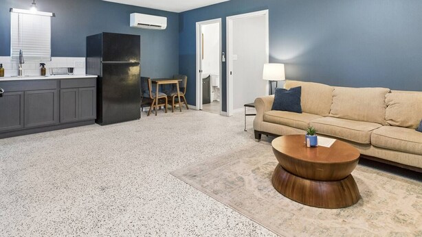 Een moderne woonkamer met een beige bank, een houten salontafel en een keukenhoek met een zwarte koelkast en witte kasten.