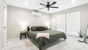Een slaapkamer met een bed, een plafondventilator, een airconditioningapparaat, een nachtkastje met een lamp en een raam met jaloezieën.