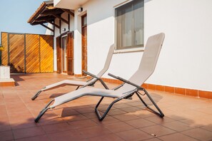 Zwei Liegestühle auf einer gefliesten Terrasse, im Hintergrund eine weiße Wand und Holzläden.