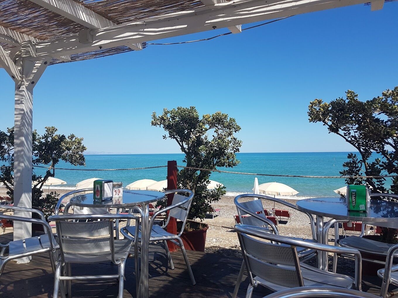 Een terras met tafels en stoelen, met uitzicht op een strand met parasols en een heldere lucht.
