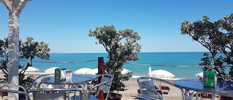 An outdoor seating area with tables and chairs, overlooking a beach with umbrellas and a clear sky.