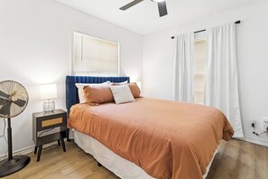 Un dormitorio con una cama, una mesita de noche, un ventilador de pie y un ventilador de techo.