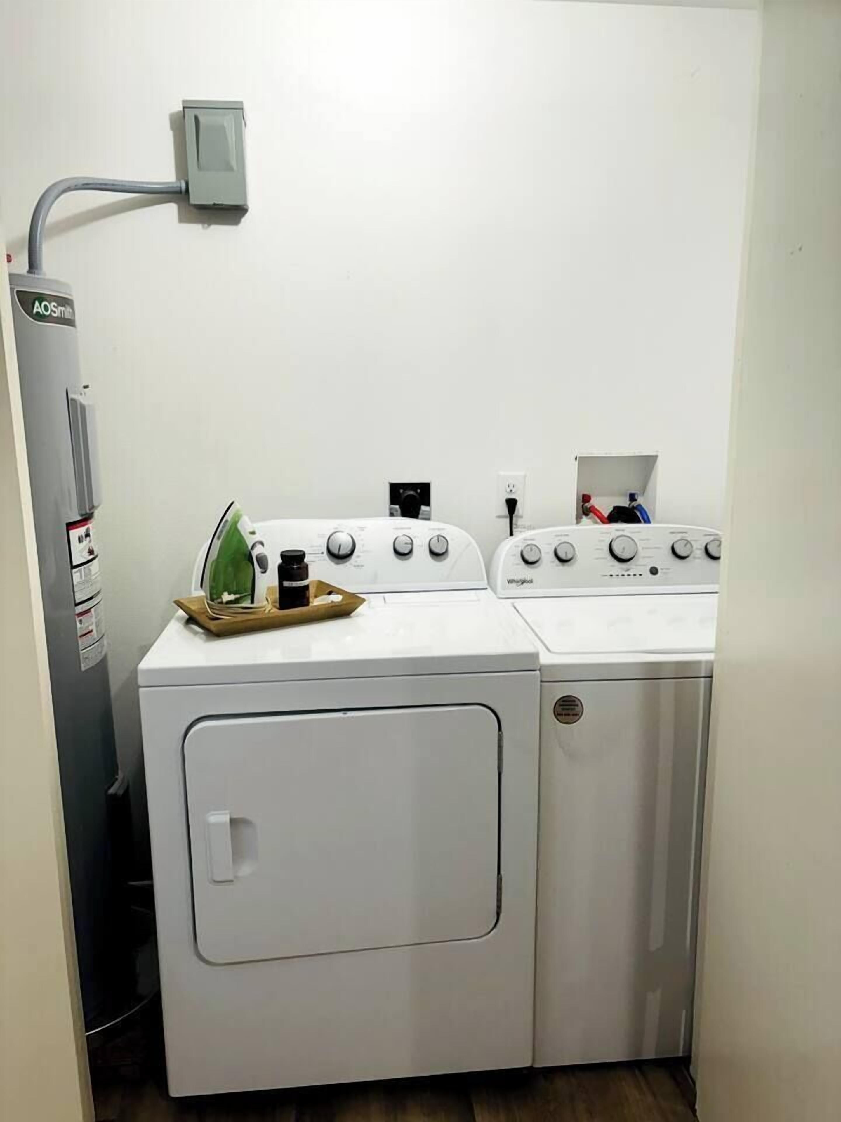 Une buanderie équipée d’un lave-linge et d’un sèche-linge à chargement frontal, d’un mur utilitaire avec une prise de courant, et d’une petite planche à repasser avec un fer à repasser et une bouteille.