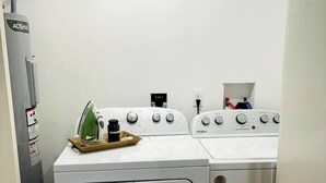 A laundry room with a front-loading washer and dryer, a utility wall with a power outlet, and a small ironing board with an iron and a bottle.