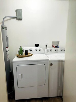 A laundry room with a front-loading washer and dryer, a utility wall with a power outlet, and a small ironing board with an iron and a bottle.