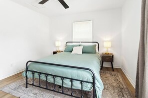 Un dormitorio con una cama, dos mesitas de noche, un ventilador de techo, una ventana con persianas y una alfombra estampada.