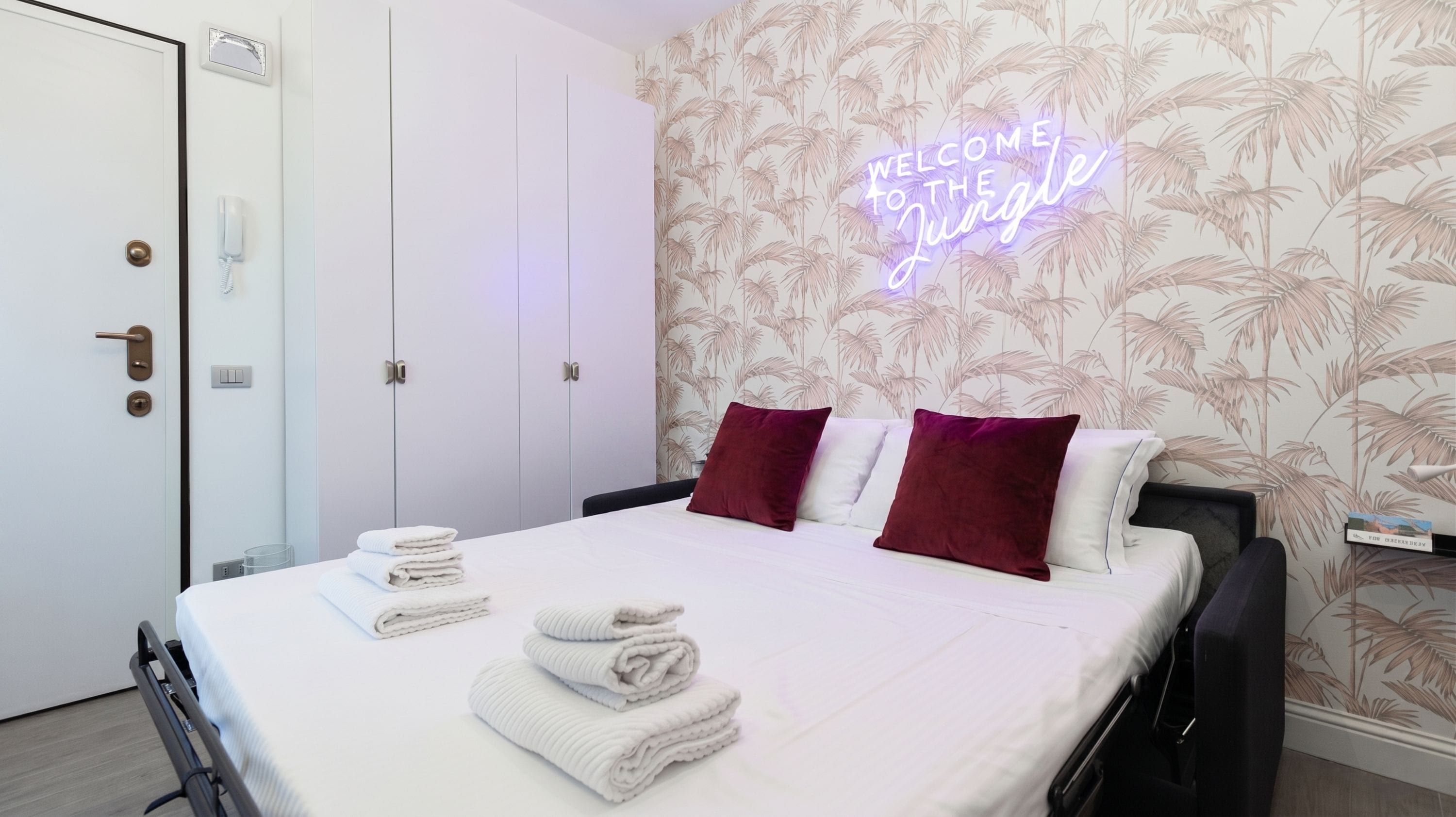 A hotel room with a bed, a neon sign, a wardrobe, and a bedside table.