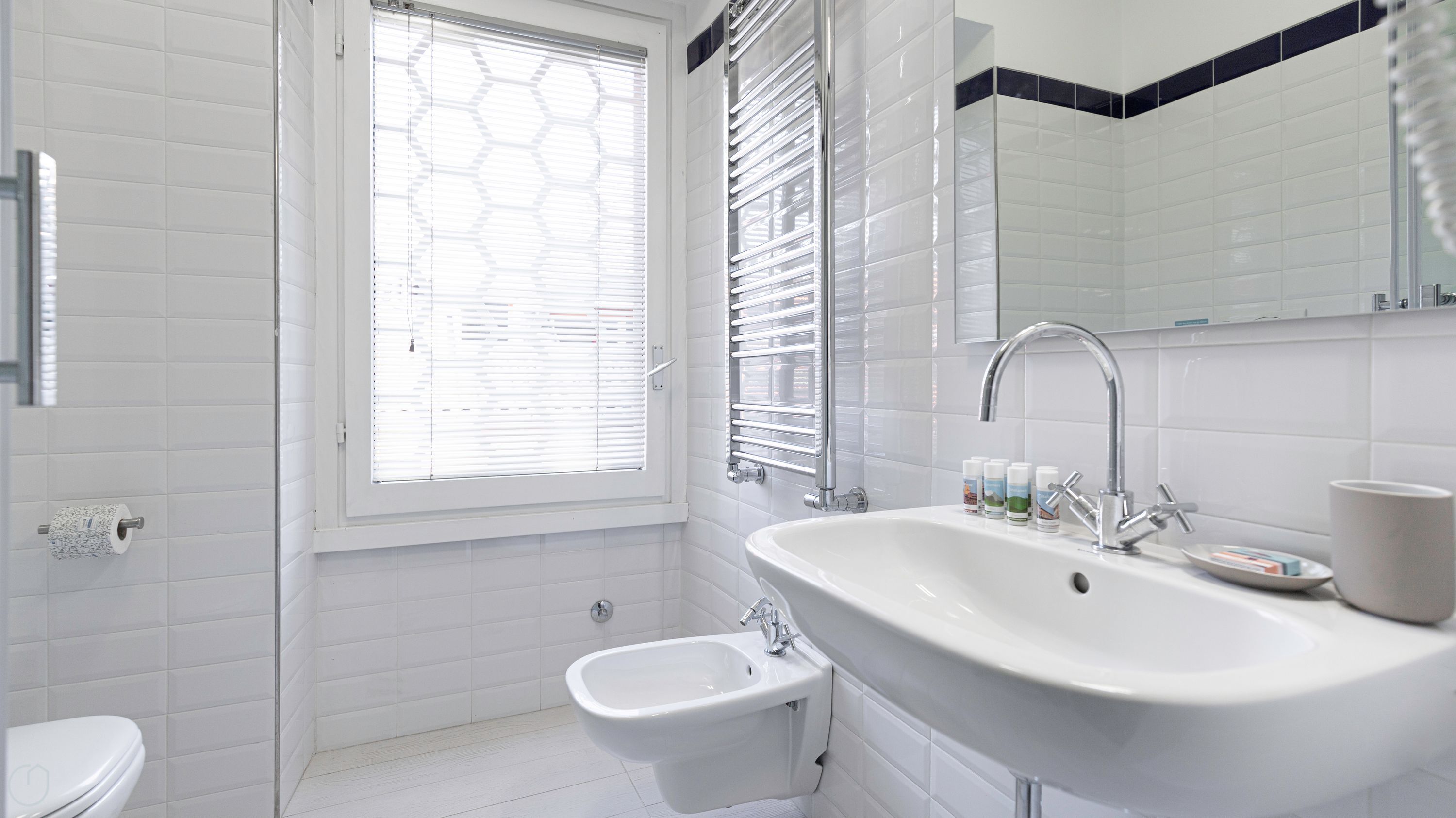 Un baño moderno con lavamanos, bidé y retrete blancos. Hay una ventana con persianas, un espejo y un perchero para toallas.