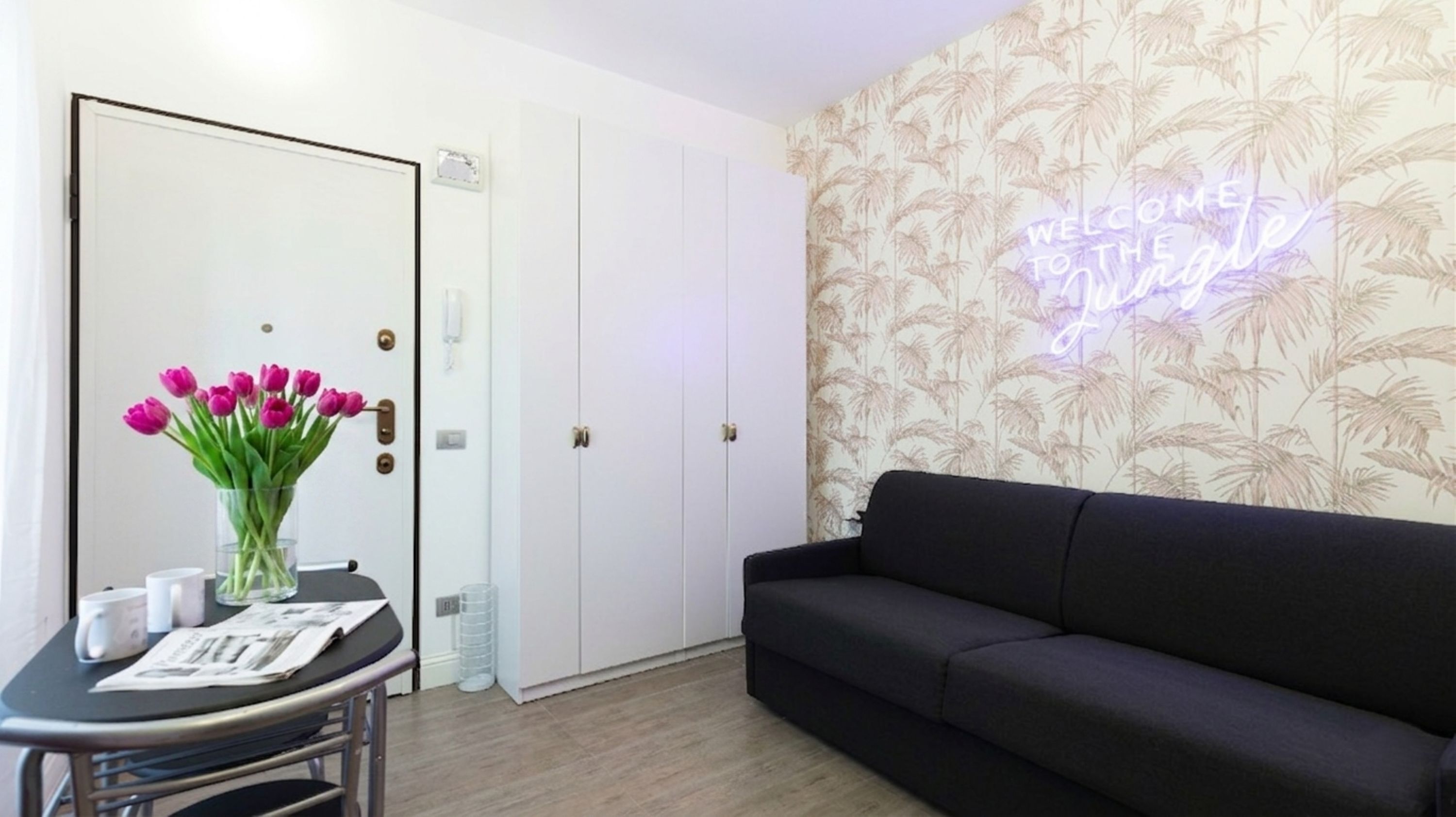 Een kamer met een gesloten deur, een zwarte bank, een tafel met een bloevas en een neonbord aan de muur.