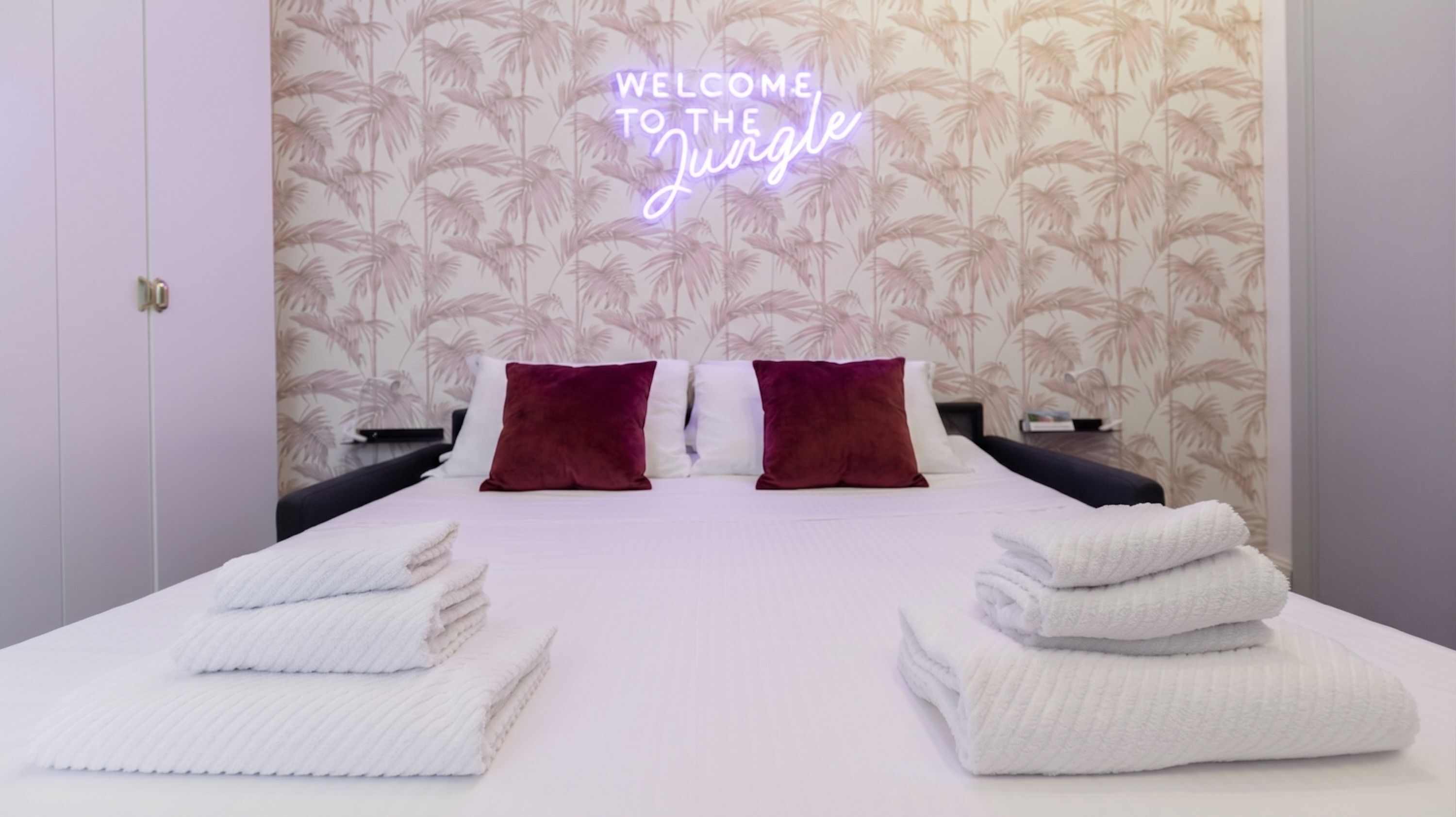 A hotel room with a bed, two pillows, a neon sign, and a tropical wallpaper.