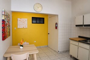 Eine Küche mit gelber Wand, einer Uhr, einem Feuerlöscher und einem Essbereich mit Tisch und Stühlen.
