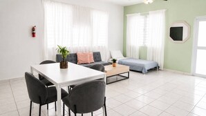 Una sala moderna con una mesa de comedor, sillas, un sofá y una cama. Hay un ventilador de techo, un extintor y una ventana con cortinas.