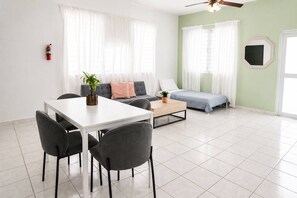 Ein modernes Wohnzimmer mit Esstisch, Stühlen, einer Couch und einem Bett. Es gibt einen Deckenventilator, einen Feuerlöscher und ein Fenster mit Vorhängen.