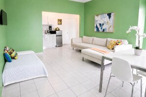 A modern living room with a sofa, a dining table with chairs, a kitchen in the background, and a green wall. - Vibrant 1BR w Balcony, Steps to Escambron Beach (San Juan)