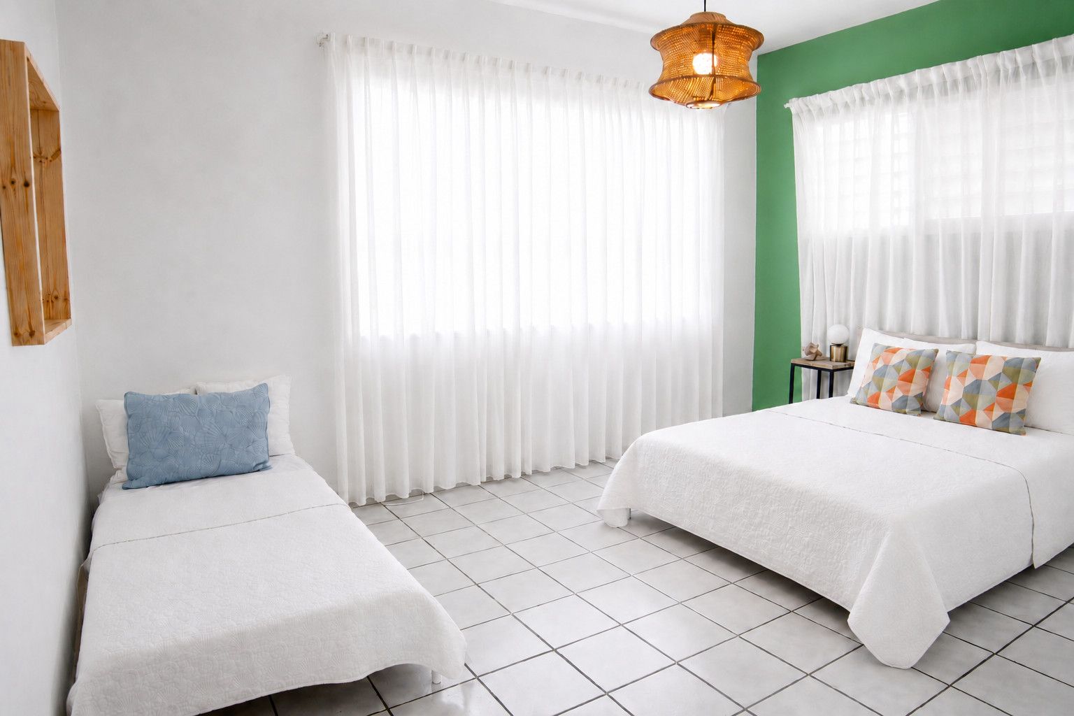 Habitación de hotel con dos camas, cortinas blancas, una pared de acento verde y una lámpara colgante.