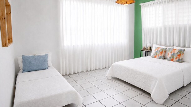 Habitació d'hotel amb dos llits, cortines blanques, una paret d'accent verd i una llum penjant.