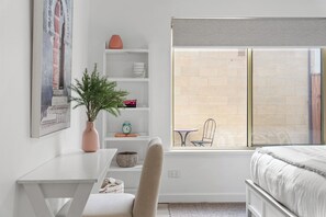 Une chambre à coucher comprenant un lit, un bureau, une chaise, une bibliothèque, une fenêtre donnant sur un mur de briques et une plante.