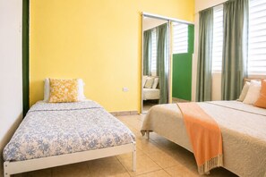A room with two beds, one with a floral bedspread and the other with a solid color bedspread. There is a green curtain and a mirror on the wall. - Charming 1 BR Walking Distance to Escambron Beach (San Juan)
