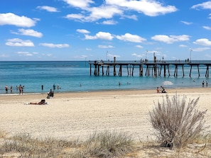 Ein Strand mit einem Steg, der sich ins Wasser erstreckt, Menschen beim Schwimmen und Sonnenbaden, und einem klaren Himmel.