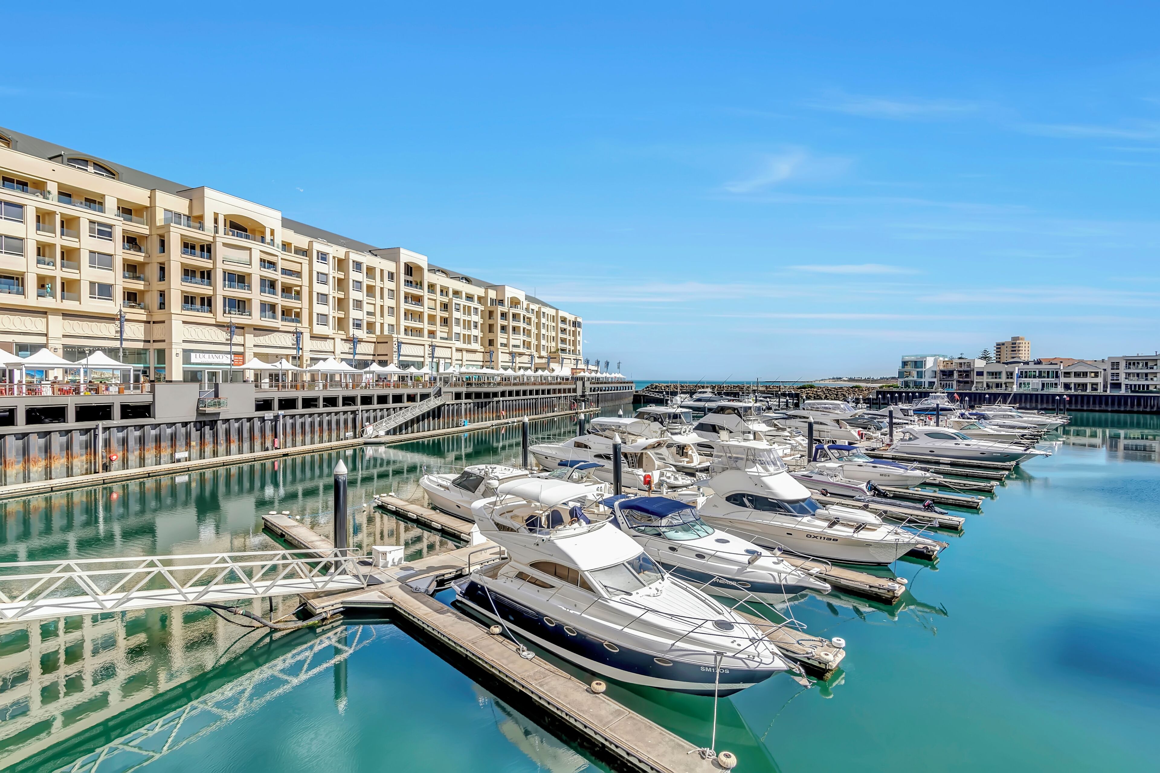 A marina with multiple boats docked, a large hotel building with balconies, and a clear blue sky.