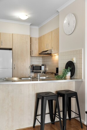 A modern kitchen with wooden cabinets, a stainless steel sink, a microwave, and a clock on the wall. - Colley Escape Vibrant Life on Glenelgs Foreshore (Glenelg)