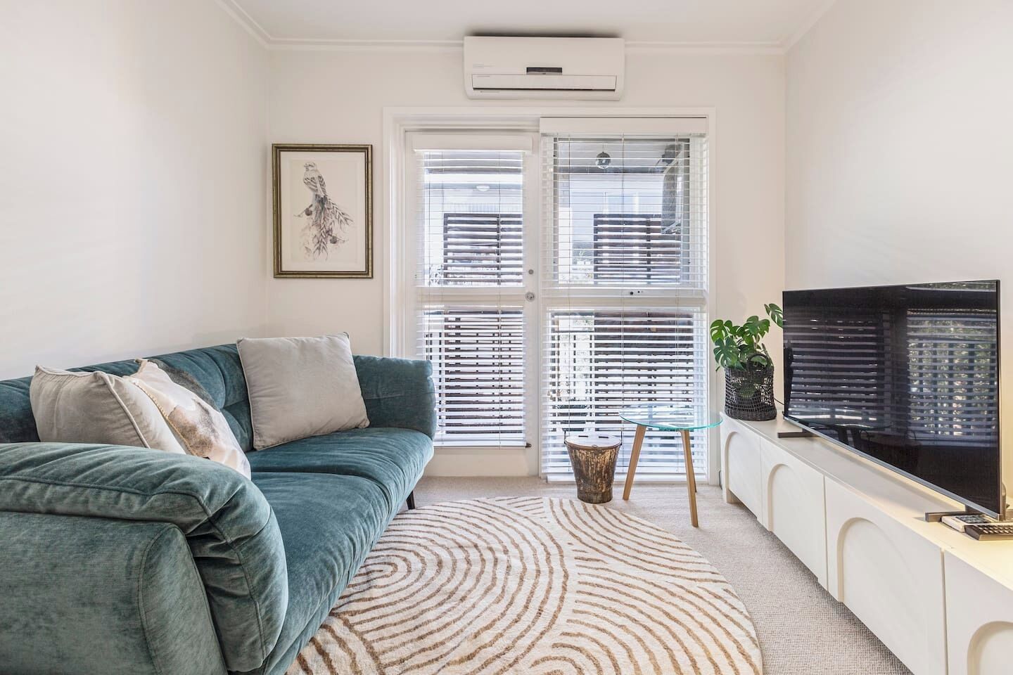 Un salon avec un canapé de couleur turquoise, un tapis à motifs, un téléviseur posé sur un meuble blanc et une fenêtre avec des stores.