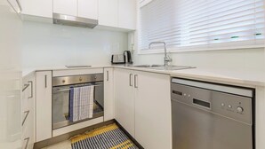 Eine kleine Küche mit weißen Schränken, einem Edelstahlbackofen, einem Geschirrspüler und einem Fenster mit Jalousien.